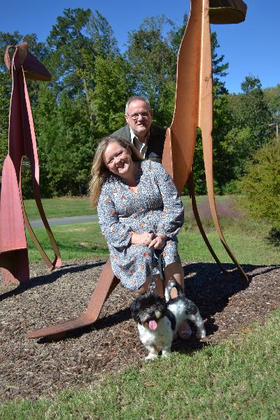 Photo of man and woman with dog posing next to "Sit & Stay" sculpture in front of shelter