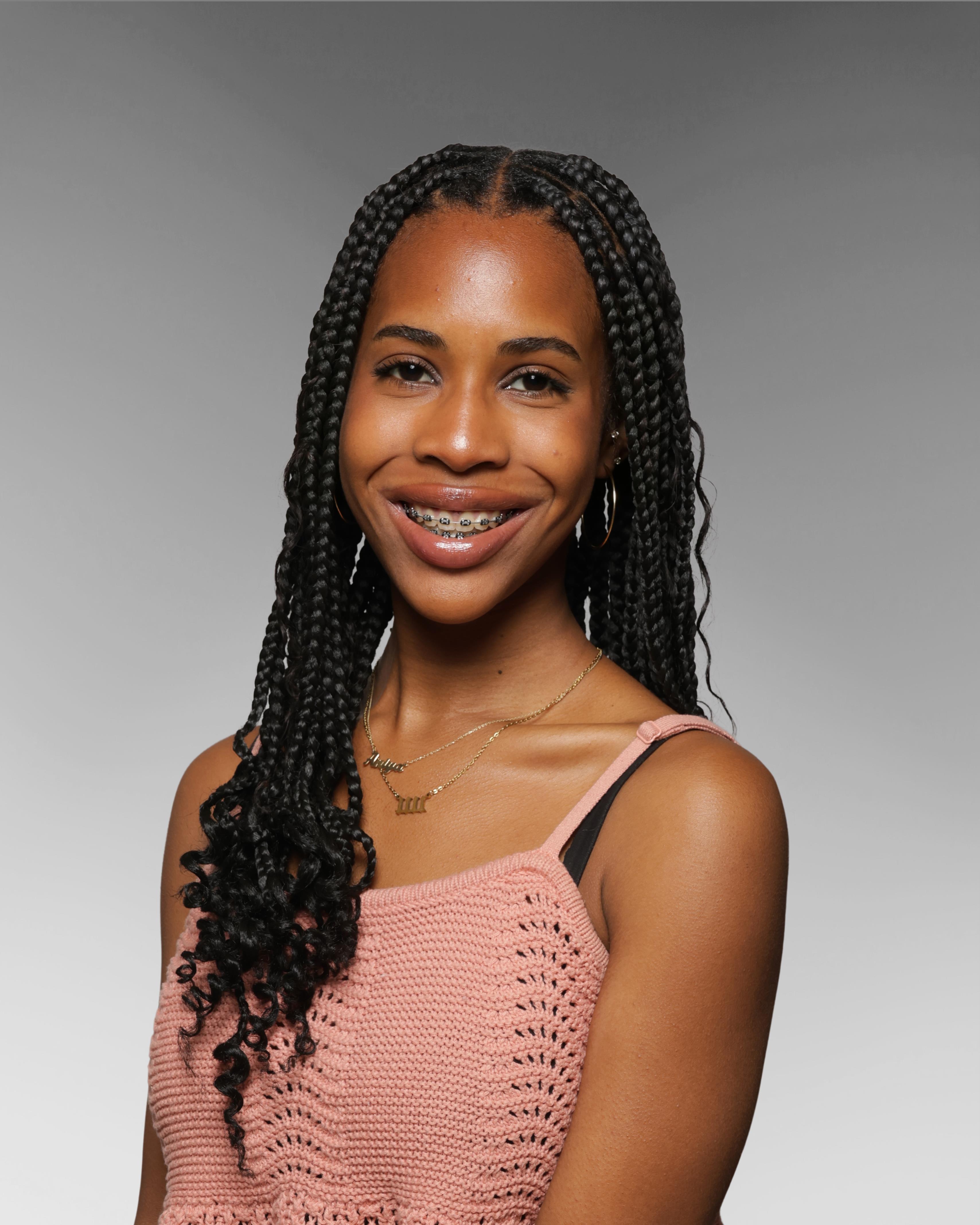 Black teen with braids, pink top, smiling against gray background.